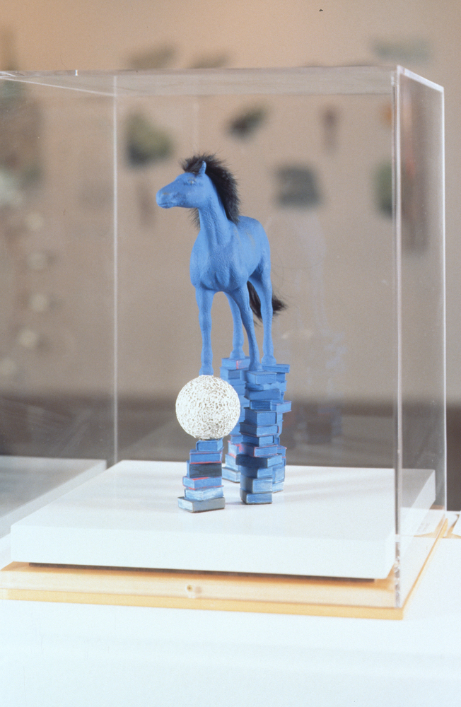 Image of a sculpture of a blue horse standing on books held within a plexiglass cover.